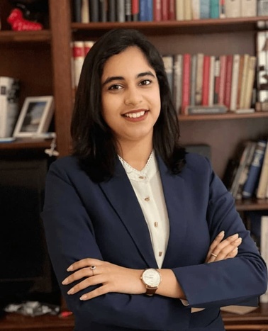 A smiling woman in a blue blazer stands confidently with arms crossed in front of a bookshelf.