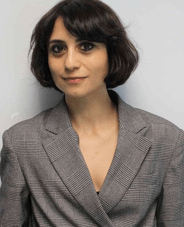 A woman with short dark hair wearing a gray blazer stands against a plain background.