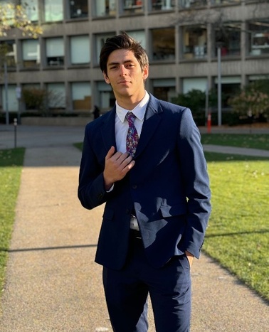 A young man in a blue suit adjusting his tie, standing outdoors with buildings in the background.