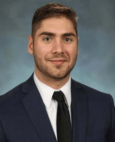 Portrait of a young man in a navy blue suit with a black tie and a light grey background.