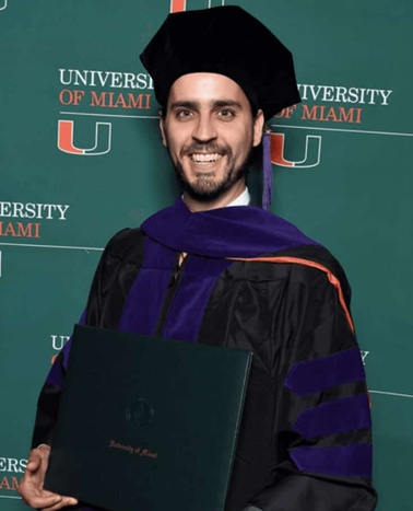 A male graduate in a black and purple academic regalia holding a diploma folder at the University of Miami.
