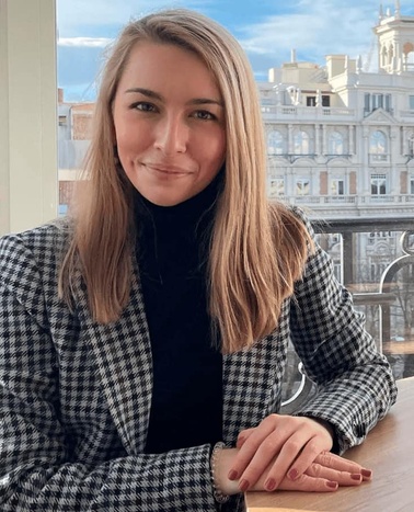 A woman in a houndstooth coat sits at a table by a window overlooking city buildings.