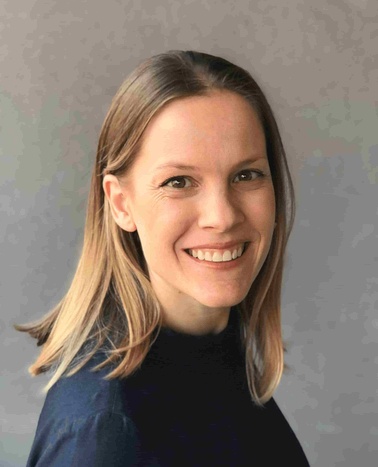 A portrait of a smiling woman with blonde hair wearing a dark blue shirt, set against a light gray background.