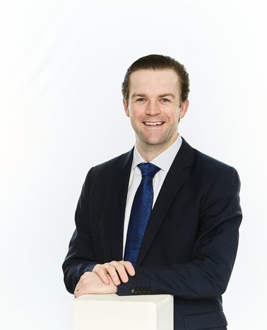 A man in a dark suit and blue tie smiling at the camera against a white background.