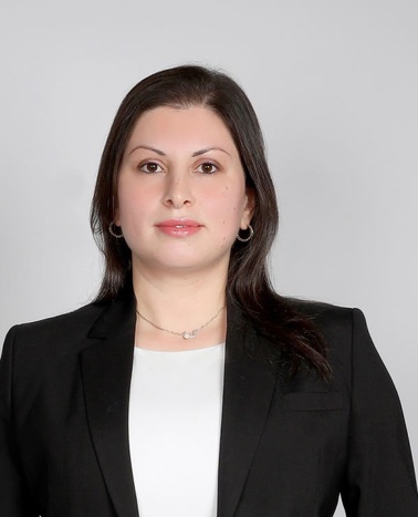 A professional portrait of a woman with medium-length hair wearing a black suit and a subtle necklace, standing against a plain light background.