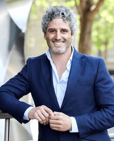 A smiling man in a blue suit standing outdoors with a casual posture.