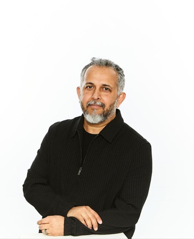 A middle-aged man with gray hair and a beard stands confidently with his arms crossed, wearing a black zip-up sweater, against a white background.