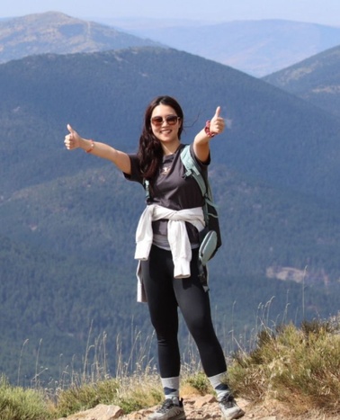 A young woman is hiking in the mountains, giving a thumbs up with a scenic backdrop.