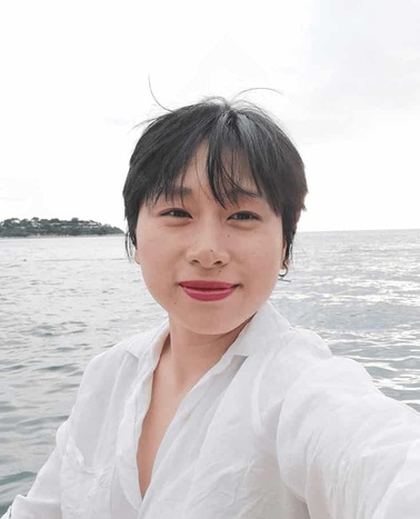 A young woman in a white blouse taking a selfie with the ocean and overcast sky in the background.