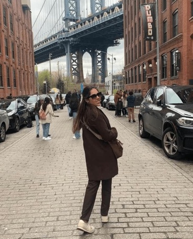 A woman poses on a street with the Brooklyn Bridge in the background, framed by red brick buildings.