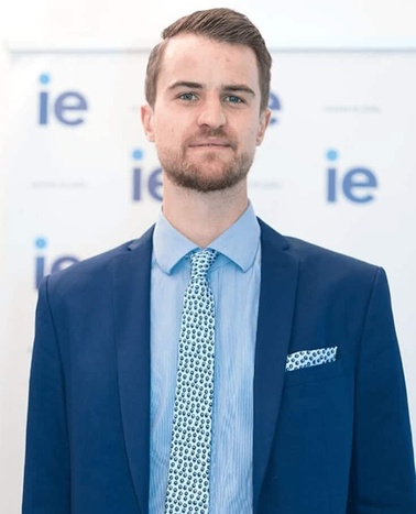 A young man in a business suit with a blue tie and a pocket square standing in front of a background with logos.