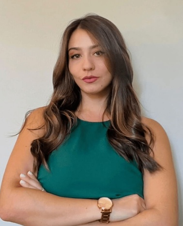 A woman in a green top with her arms crossed stands against a light-colored background.
