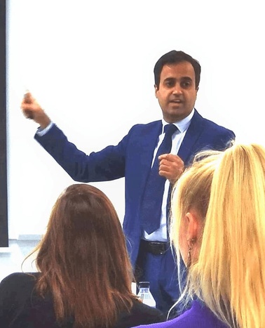 A man in a blue suit is presenting in front of an audience, pointing to his right.