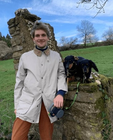 A young man in a light coat smiling next to two dogs on a stone structure in a green field.