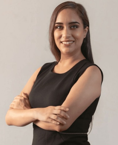 A confident young woman in a black sleeveless dress, smiling and standing with her arms crossed.