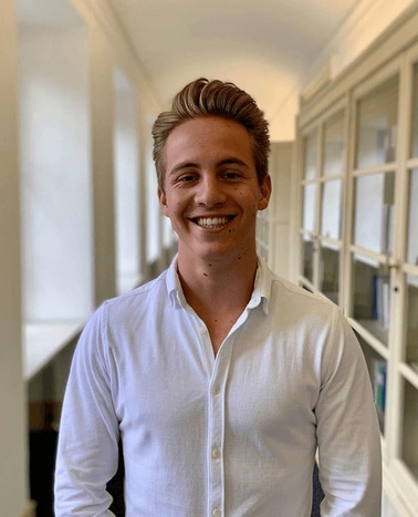 A smiling young man in a white shirt standing in a hallway with windows.