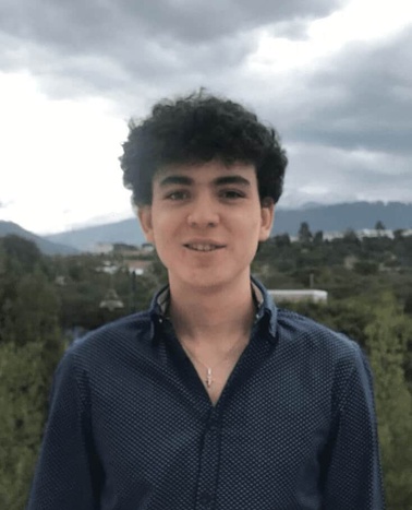 A young person with curly hair wearing a blue shirt standing outdoors with a blurred landscape of trees and mountains in the background.