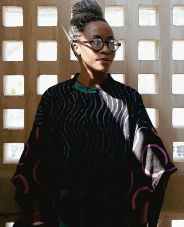 A woman wears a stylish black and colorful garment while standing in front of a textured wall.