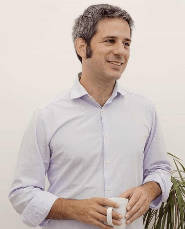 A man in a light blue shirt holding a white mug, smiling and looking to the side.