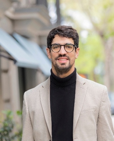 A man in glasses, wearing a beige blazer and black turtleneck, smiles while standing on a city street.