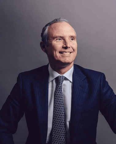 A portrait of an elderly Caucasian man smiling, dressed in a dark blue suit with a tie, posed against a neutral background.