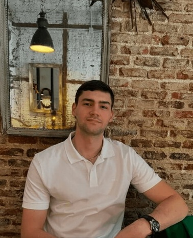A young man sitting in front of a brick wall with a lamp in a frame behind him