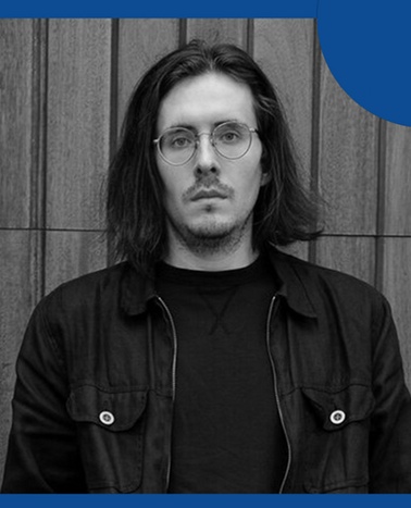 A young man with long hair wearing glasses and a black jacket stands against a wooden panel background.