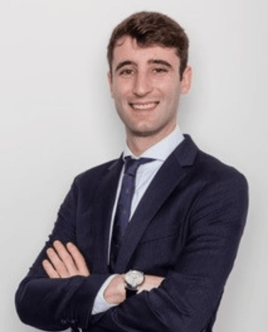 A young man in a business suit smiling with crossed arms against a white background.