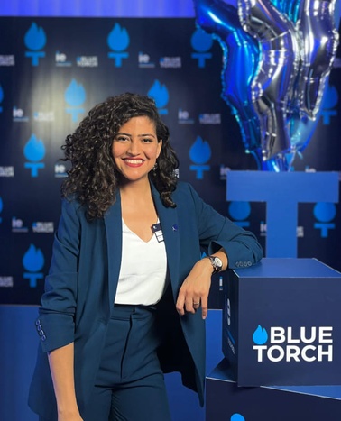 A woman in a blue suit is smiling while standing at a podium with the 'Blue Torch' logo.