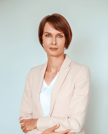 A professional looking woman with short hair stands confidently with her arms crossed, wearing a light pink blazer over a white blouse against a pale blue background.