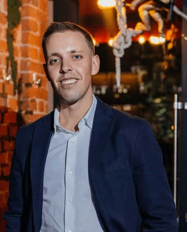 A smiling man in a suit standing in a bar with brick walls and decorative lights.