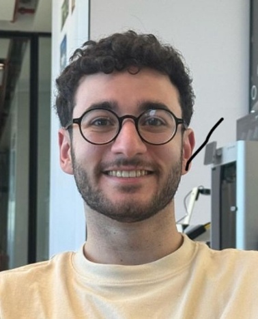 A young man with curly hair and glasses, smiling in an indoor environment.