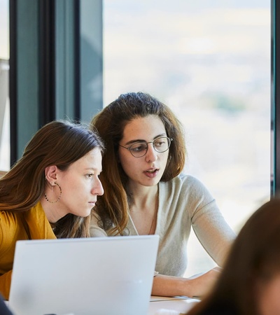 Two women collaborate while working on laptops in a bright, modern workspace.