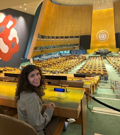 A young woman sits at a desk in the United Nations General Assembly hall, surrounded by rows of empty seats.