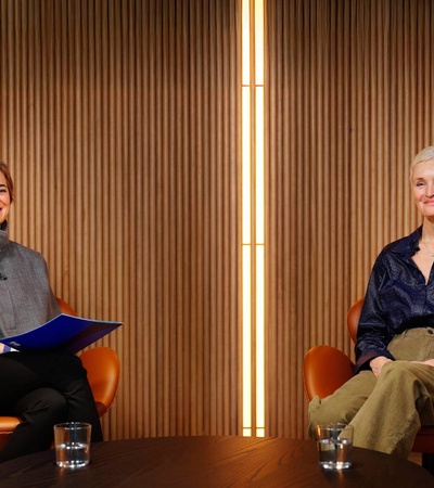 Two women seated in stylish chairs engage in conversation on a modern set.