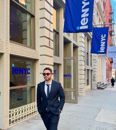 A man in a suit walking in front of a college building with flags displaying its name.