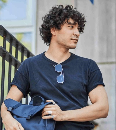 A young man with curly hair standing outdoors holding a jacket over his shoulder.