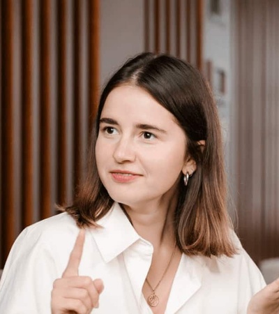 A young woman in a white blouse is gesturing with one hand raised, looking as though she is explaining something and smiling subtly, in an office setting with a wooden backdrop.