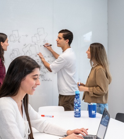 A group of four young people collaborate in a modern workspace, with one drawing on a whiteboard while others engage in discussion.