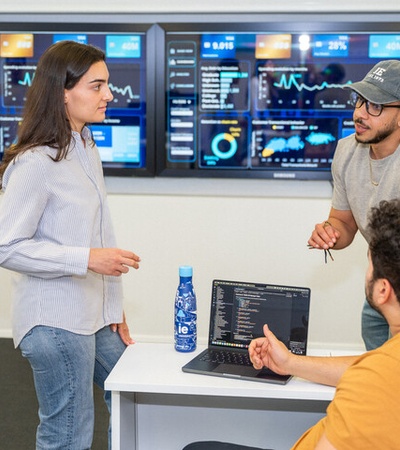 A group of three people having a discussion in an office setting with digital screens displaying data in the background.