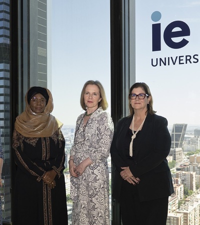 Five women standing in a modern office setting with large windows overlooking a cityscape, next to a sign that reads 'IE University'.