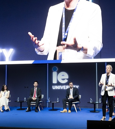 A professional conference event with speakers presenting to an audience, featuring a large screen displaying a close-up of a speaker.