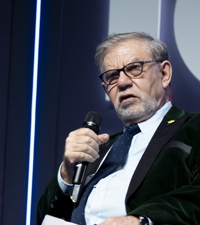 An elderly man with glasses speaking into a microphone at a conference, with a logo visible in the background.