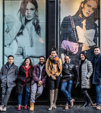 A group of seven people standing in front of a store with large fashion advertisements in the background.