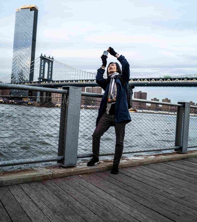 A person is taking a picture with a smartphone by a river with a bridge in the background.