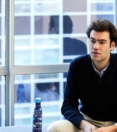 A man sitting on a desk in a well-lit room with large windows in the background.