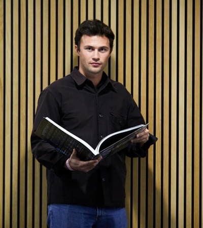 A man stands holding a book open in front of a yellow striped wall.