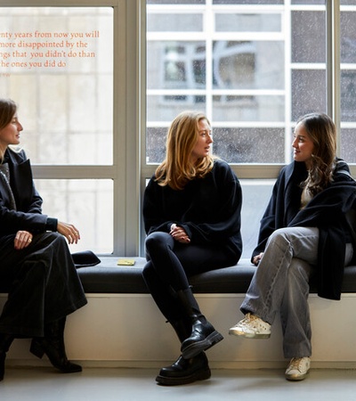 Three women in dark coats sitting and chatting by a large window in a room.