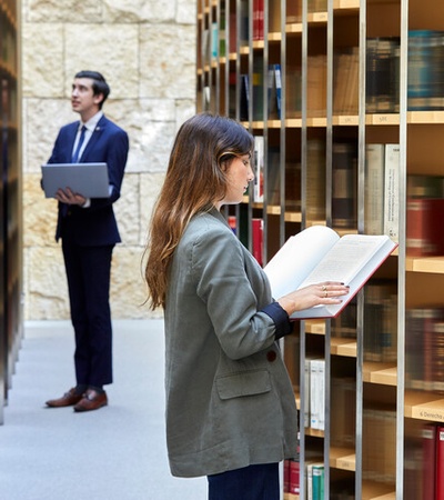 A woman and a man, dressed in business attire, are reading books in a library with floor-to-ceiling shelves filled with books.