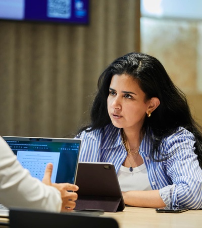 A woman engages in a serious discussion at a meeting table, while a man is seated across from her with a laptop.